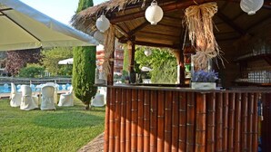 Estrutura de bambu com lanternas penduradas, uma piscina e cadeiras brancas em um jardim.