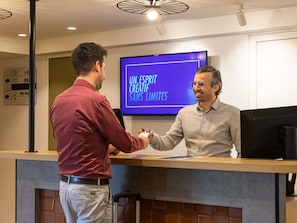 A reception desk with a flat-screen TV displaying 'UN. ESPRIT CREATIVE SANS LIMITES' in the background. - Ibis Styles Lille Villeneuve d’Ascq (Villeneuve-d'Ascq)