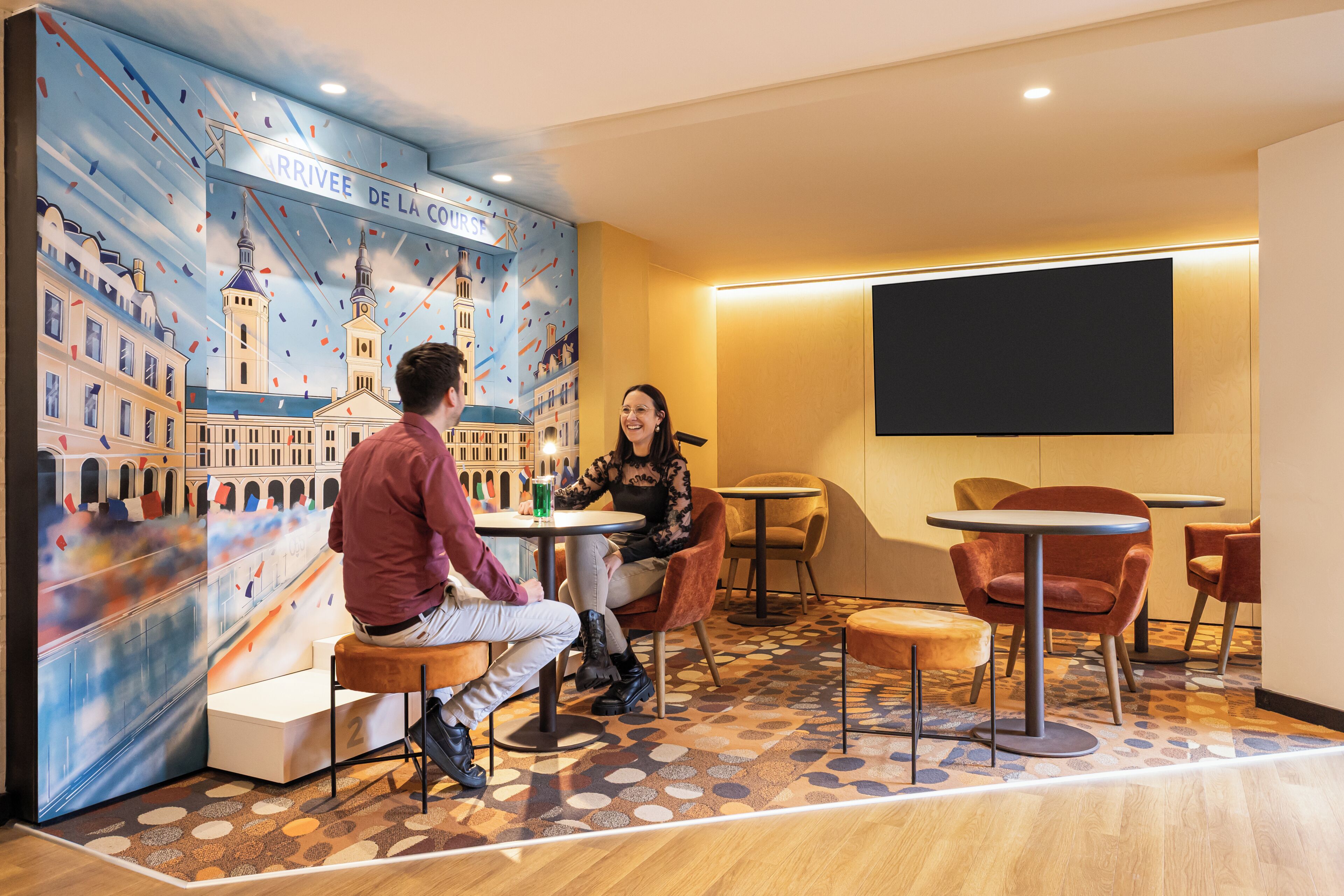 Um interior moderno com um mural de paisagem urbana, uma TV de tela plana e uma área de estar com mesas e cadeiras.