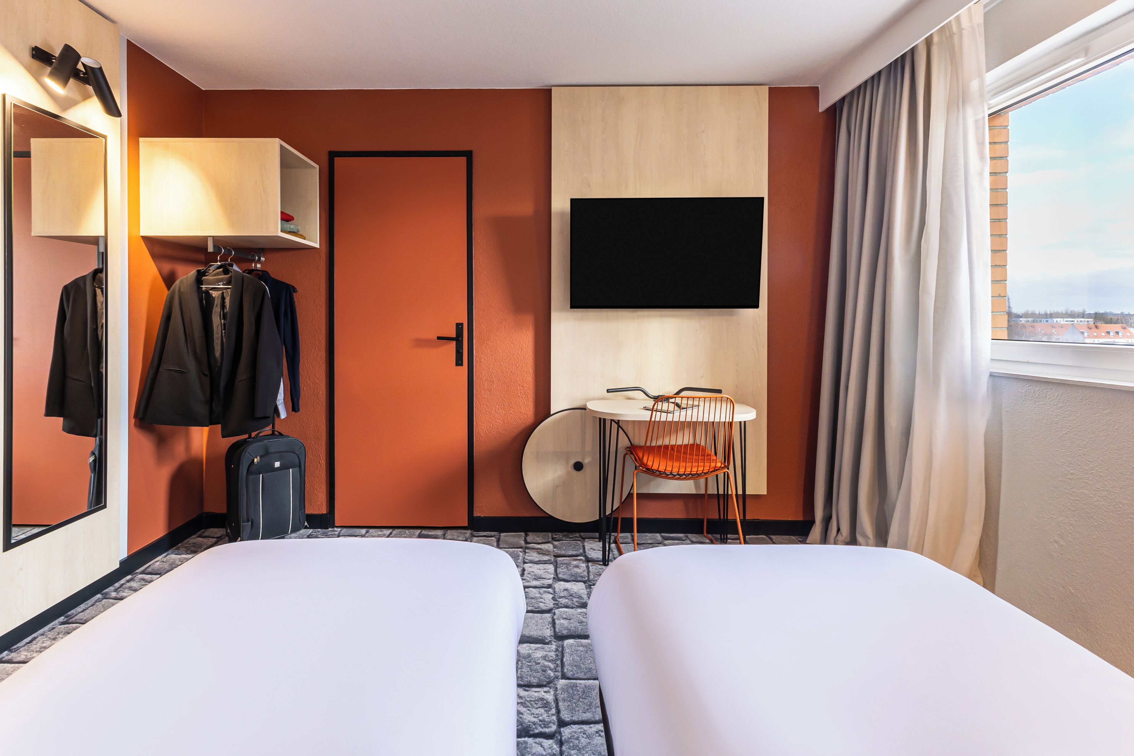 Camera d'albergo con due letti, una scrivania con sedia, una TV, uno specchio, un armadio e una valigia.
