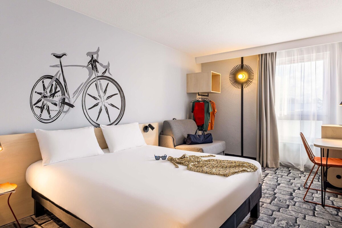 Habitación de hotel con una cama grande, un adhesivo con forma de bicicleta en la pared, un escritorio con lámpara y vistas al exterior a través de la ventana.