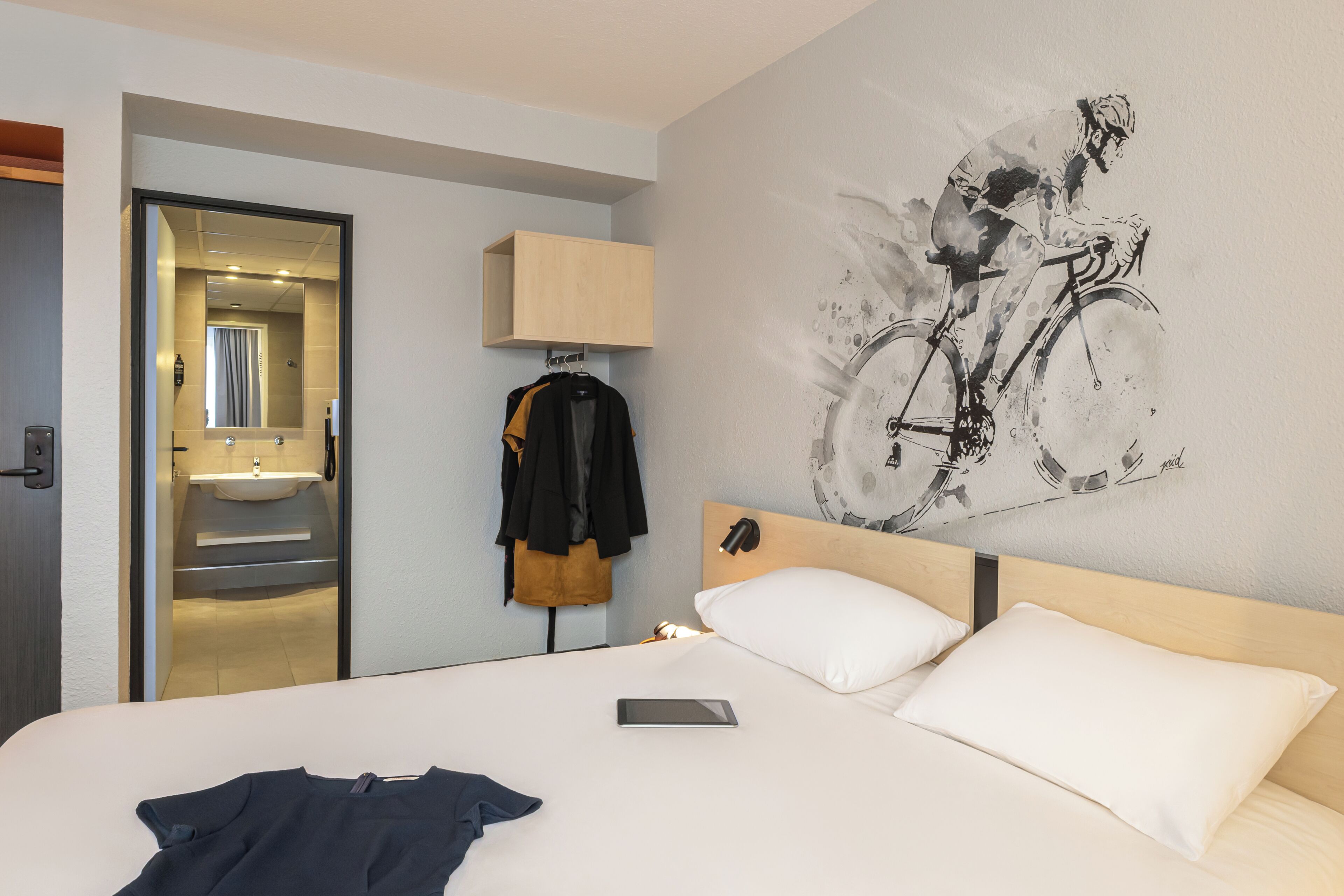 Camera d'albergo con un letto, un comodino, una lampada a parete, uno specchio, un bagno parzialmente visibile attraverso una porta aperta e un quadro raffigurante un ciclista.