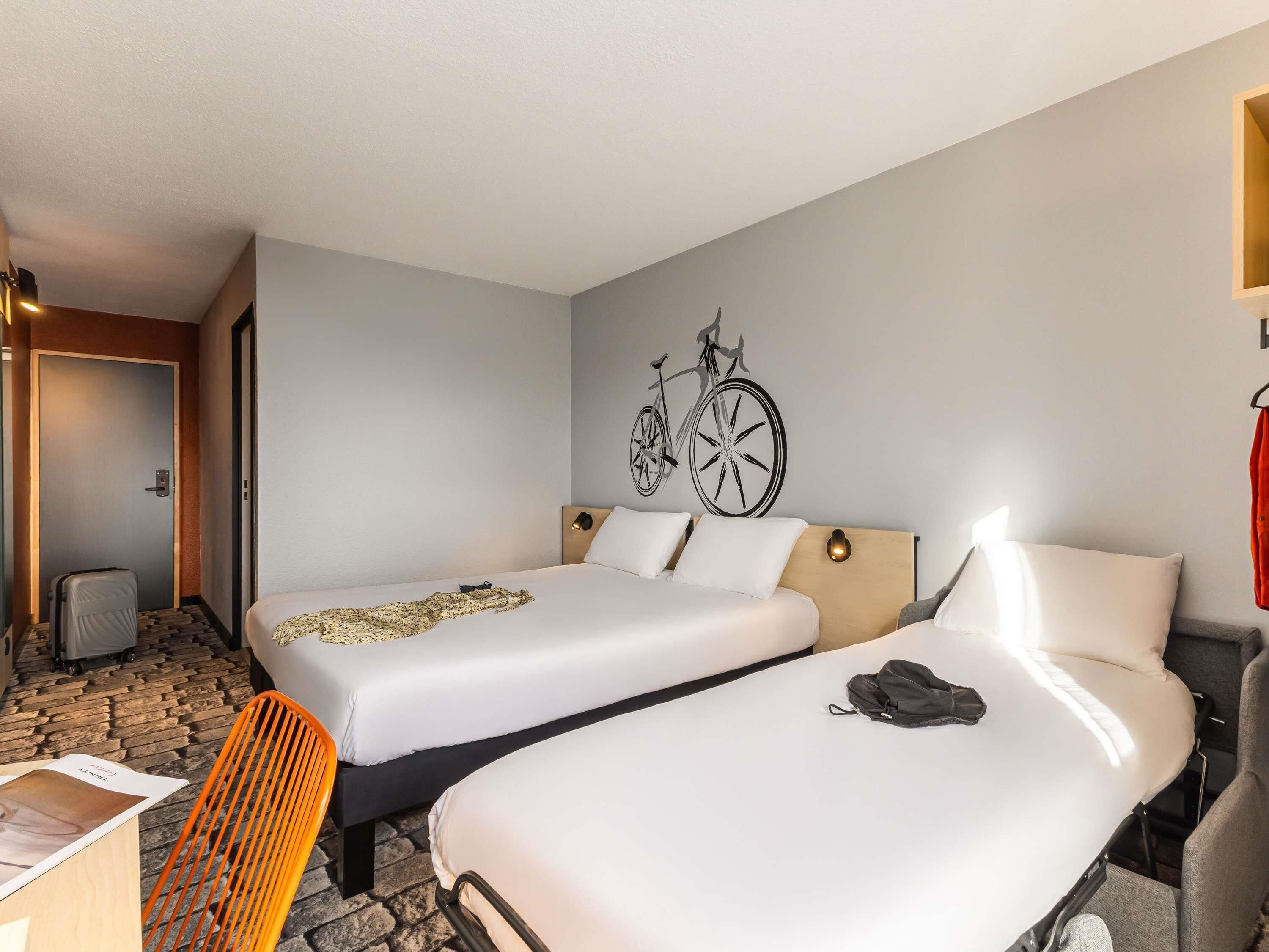 Une chambre d’hôtel avec deux lits, une œuvre d’art représentant un vélo accrochée au mur, un bureau avec une chaise et une porte donnant sur une autre pièce.