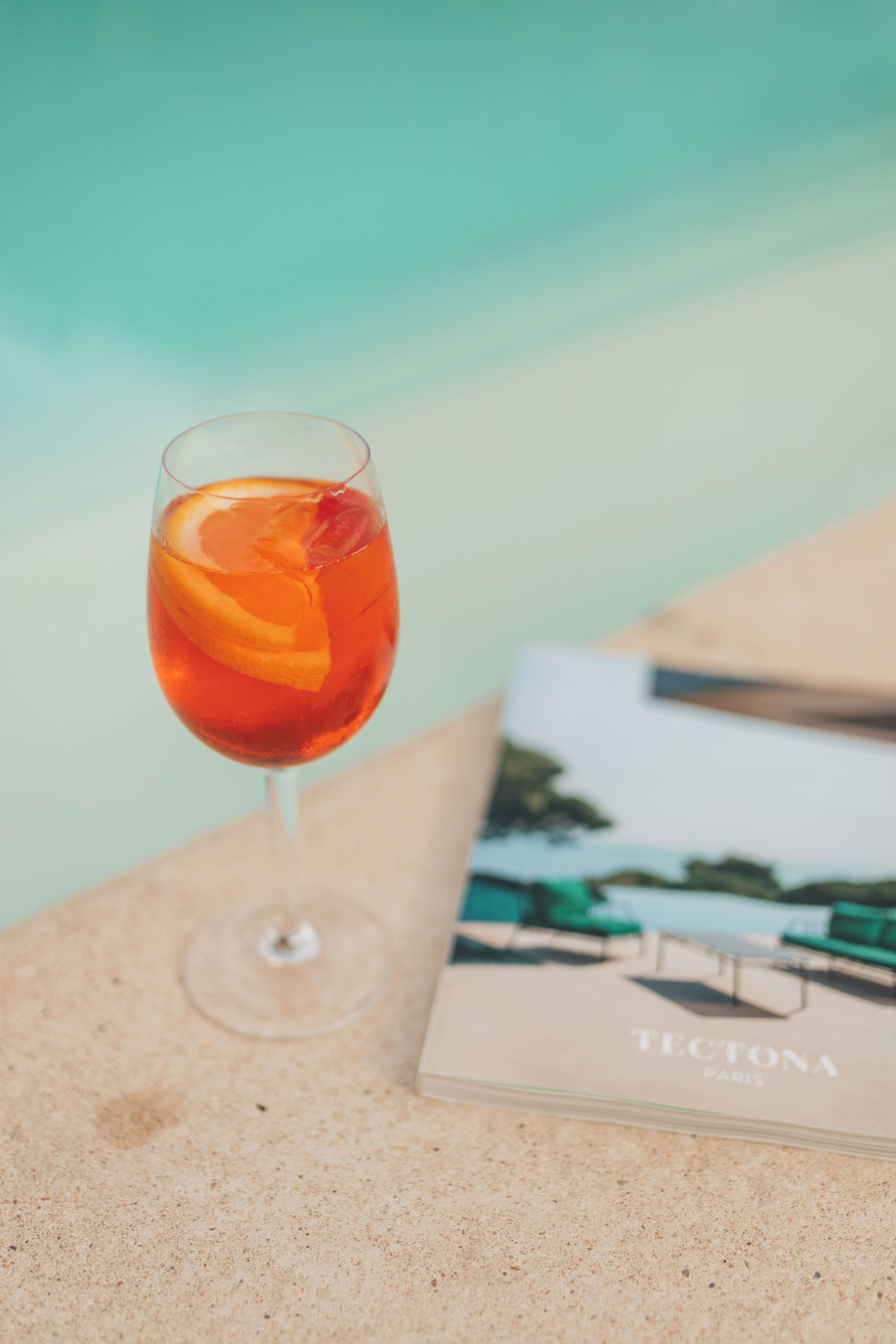 A glass of red beverage with an orange slice on a textured surface, a book titled "TECTONA PARIS" beside it.