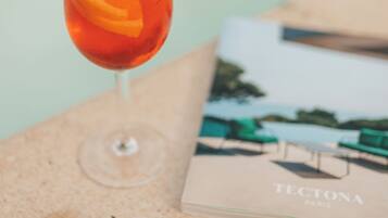 A glass of red beverage with an orange slice on a textured surface, a book titled "TECTONA PARIS" beside it.