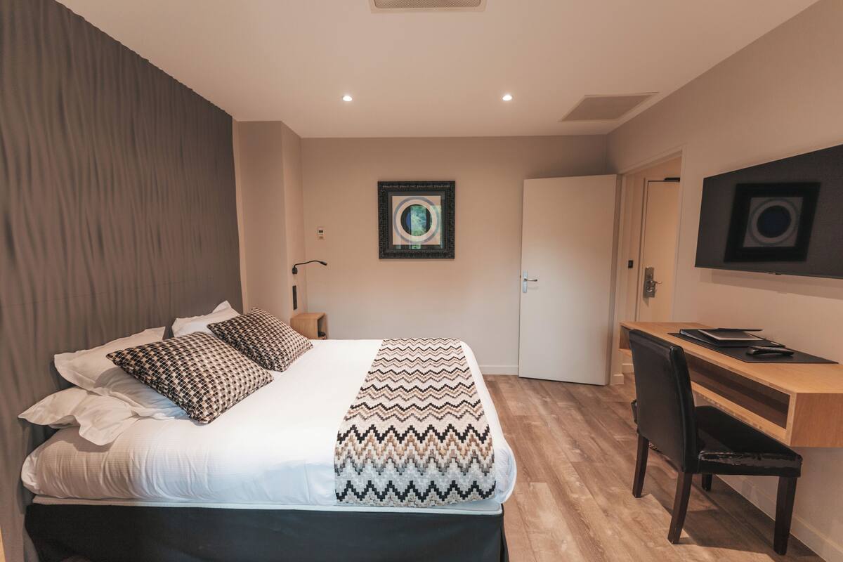 A bedroom with a bed, a desk, and a chair. There is a door and a wall-mounted picture.