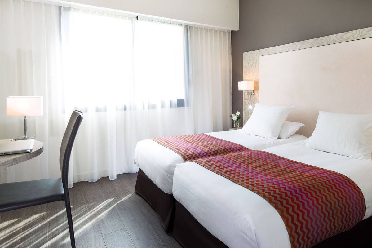 A hotel room with two beds, a desk with a lamp, and a chair.