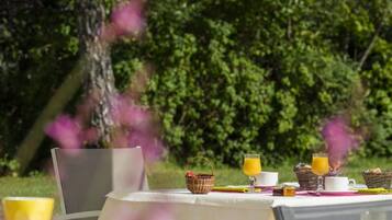A dining table set outdoors with a white tablecloth, surrounded by chairs, and various breakfast items including a basket, cups, and a glass of orange juice.