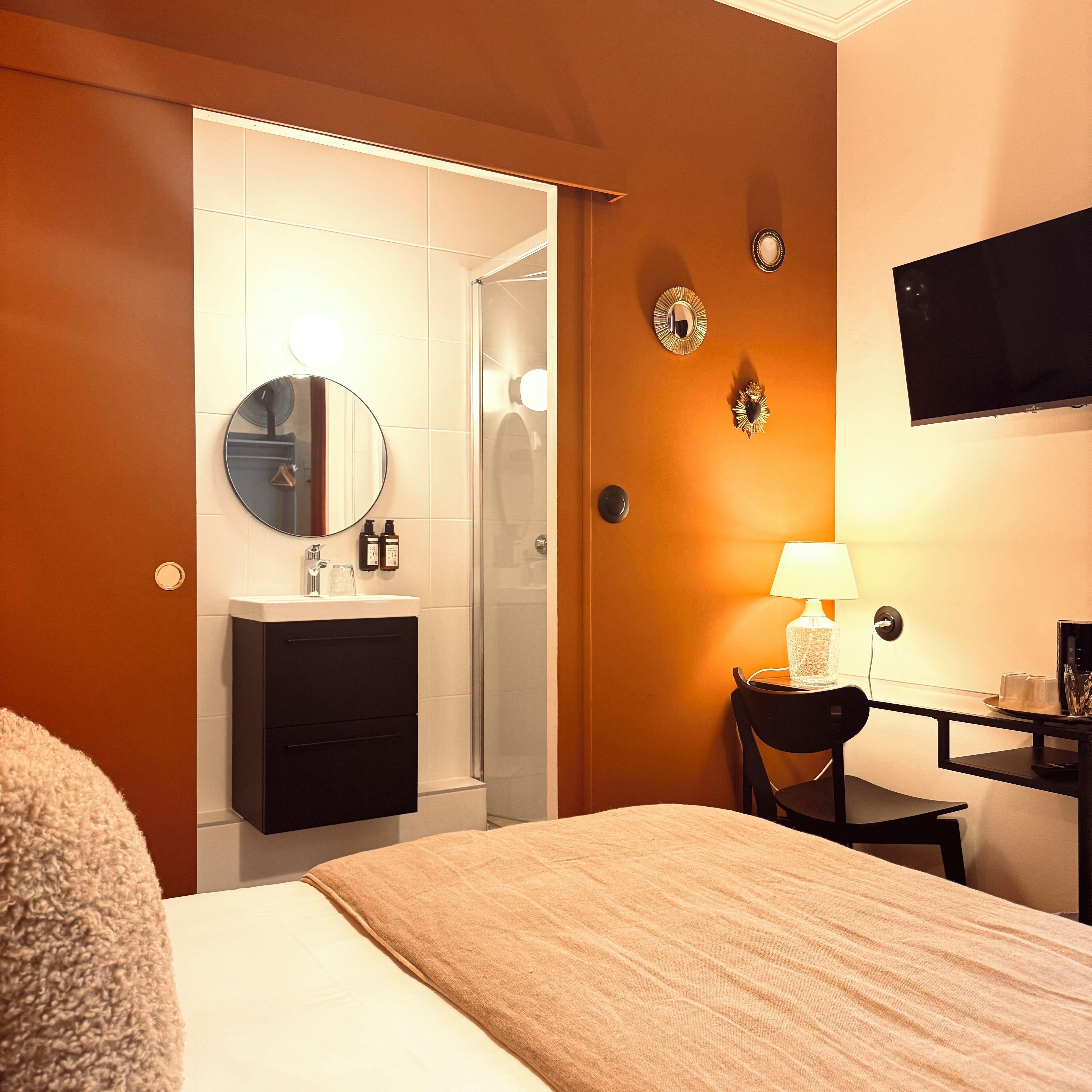 Habitación de hotel con cama, escritorio con lámpara, silla, baño con lavamanos y espejo, y televisor montado en la pared.
