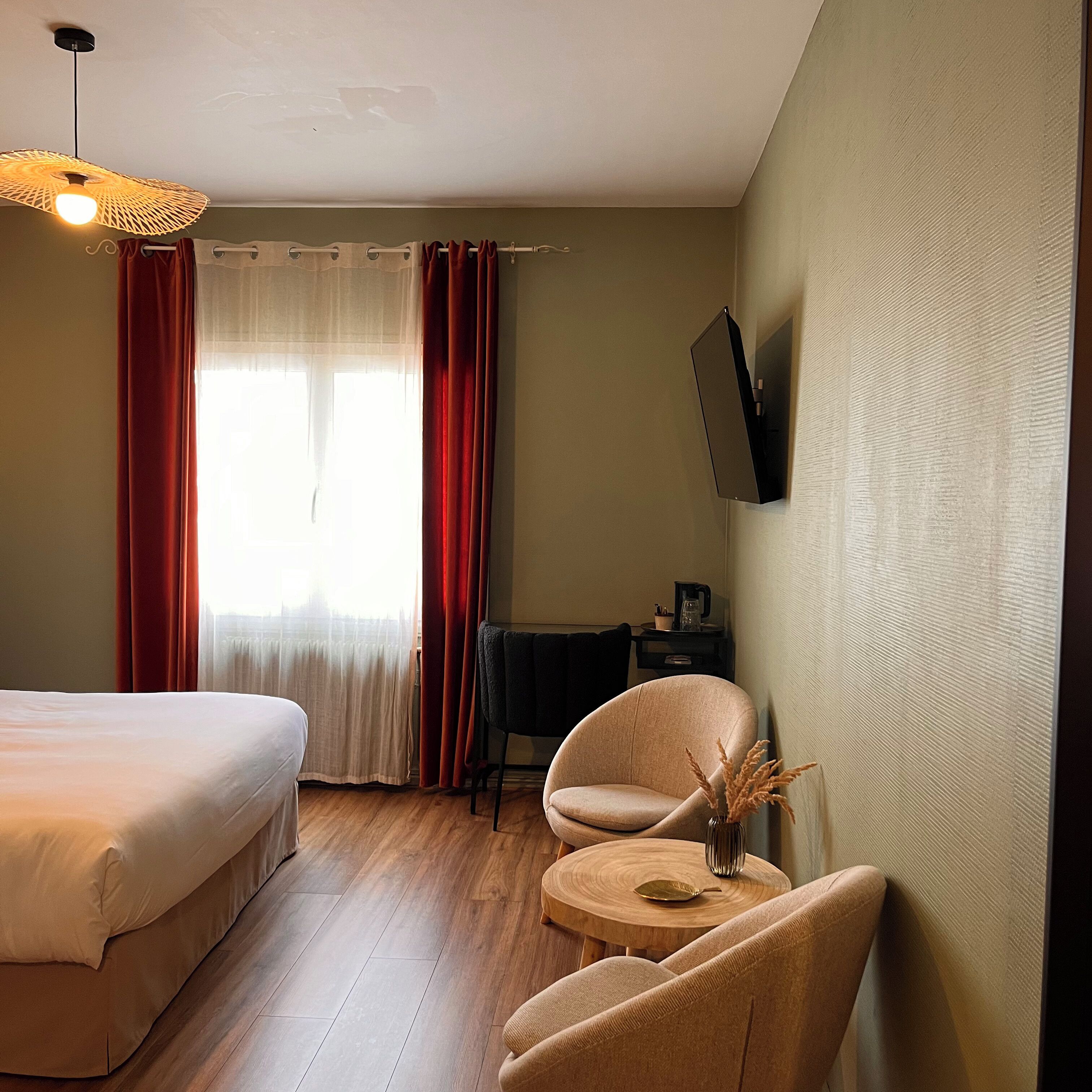 Une chambre d’hôtel comprenant un lit, deux chaises, une petite table, un téléviseur fixé au mur et une fenêtre avec des rideaux.