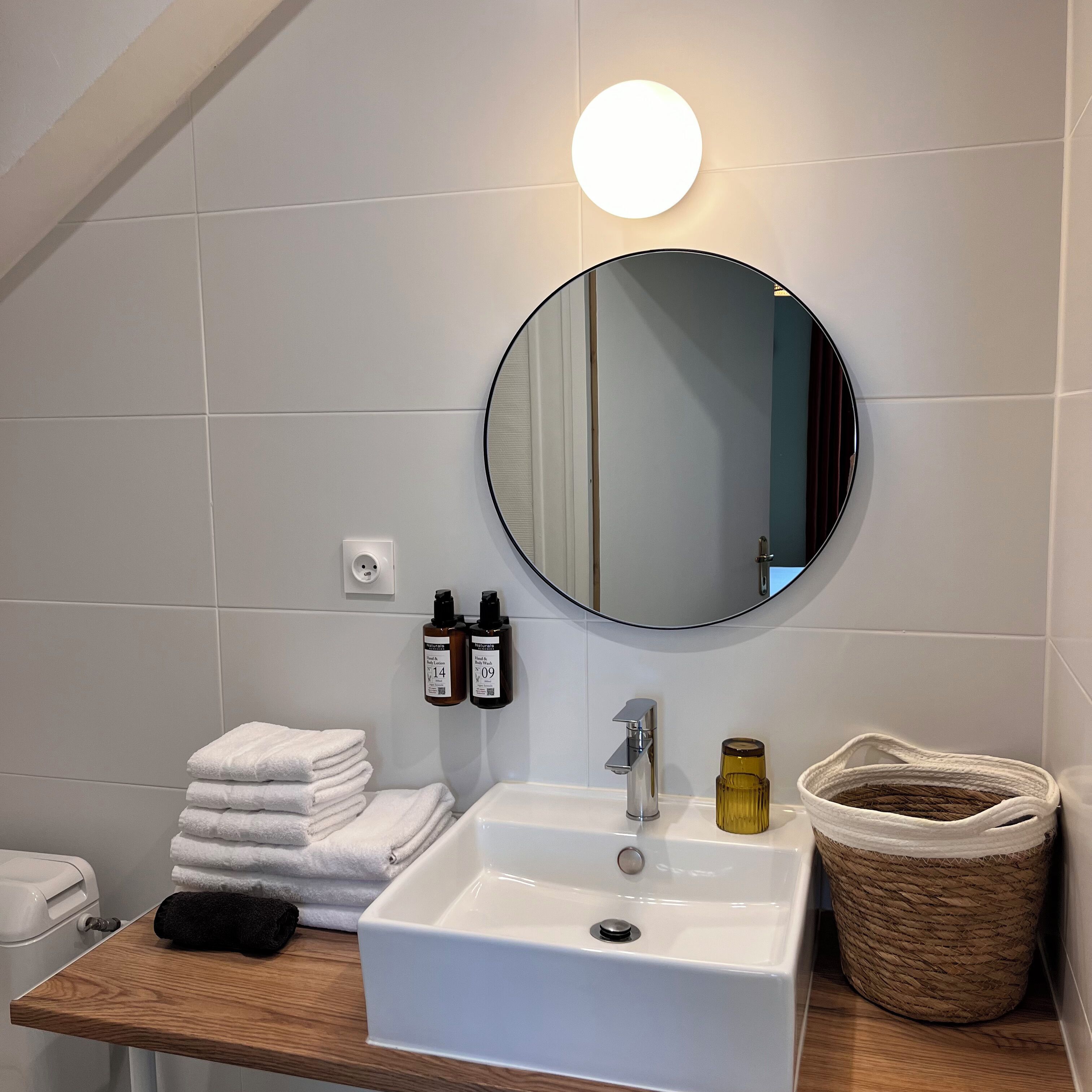 Une salle de bain avec un lavabo blanc, un miroir rond, deux bouteilles de shampoing, une pile de serviettes, une serviette noire, une bouteille jaune et un panier en osier.
