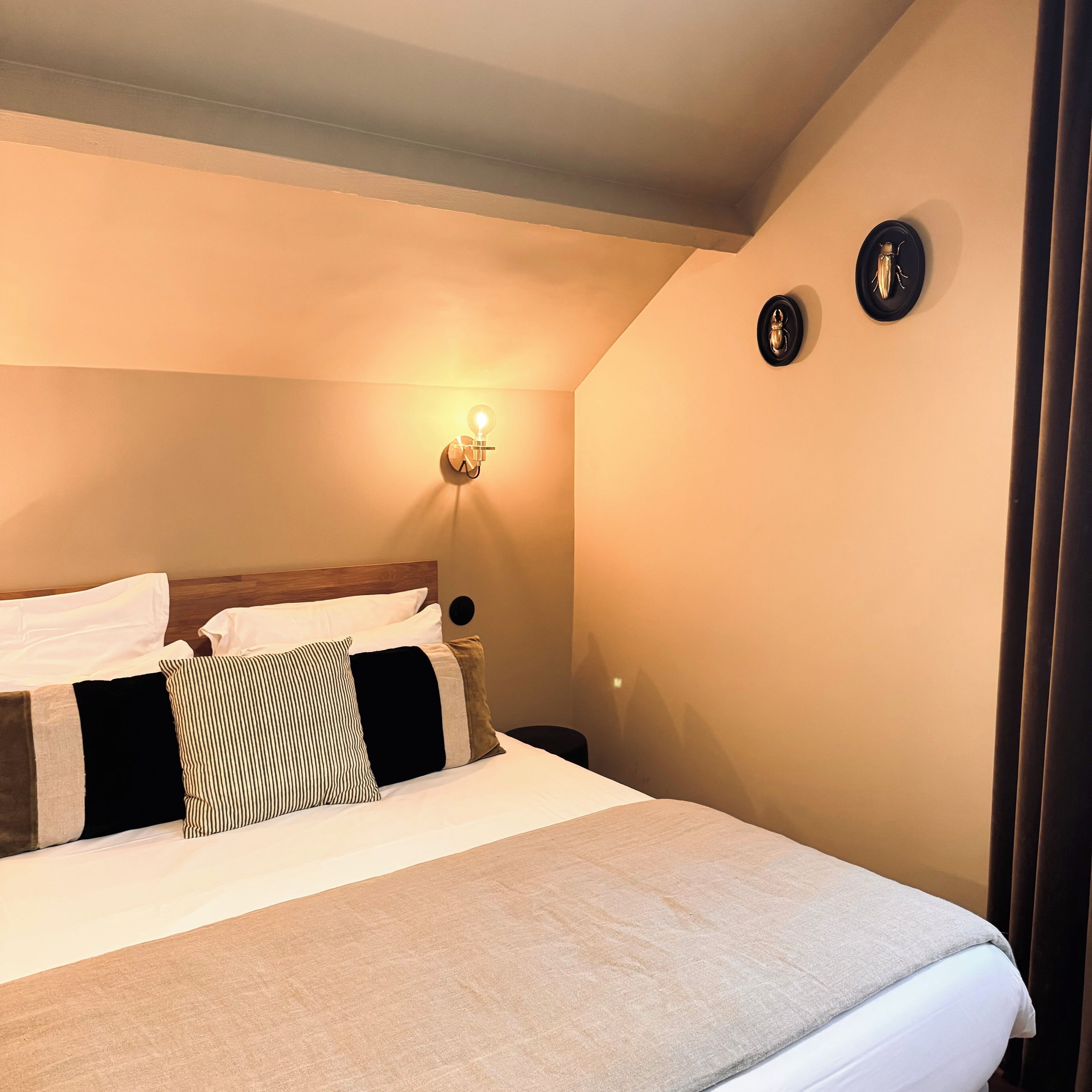 Una cama bien hecha con una manta gris y varios almohadones, una lámpara de pared y dos relojes de pared.
