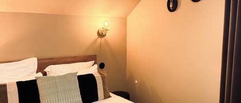 Una cama bien hecha con una manta gris y varios almohadones, una lámpara de pared y dos relojes de pared.