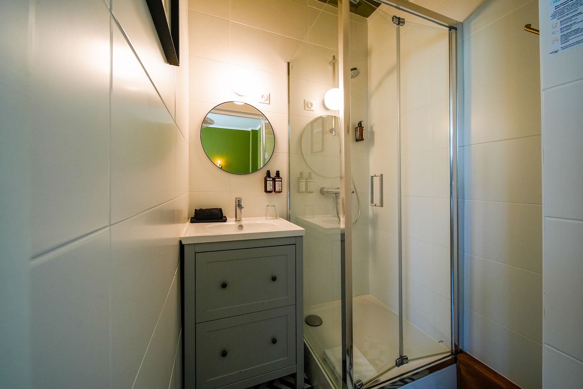 Une salle de bain moderne avec une douche à l’italienne en verre, un miroir rond et un meuble-lavabo blanc doté d’un évier et de deux tiroirs.