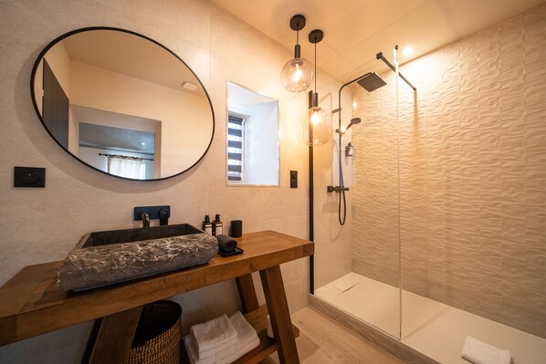 Une salle de bain avec un miroir rond, un meuble-lavabo en bois, un vasque en pierre, une douche avec un mur texturé et des luminaires suspendus.