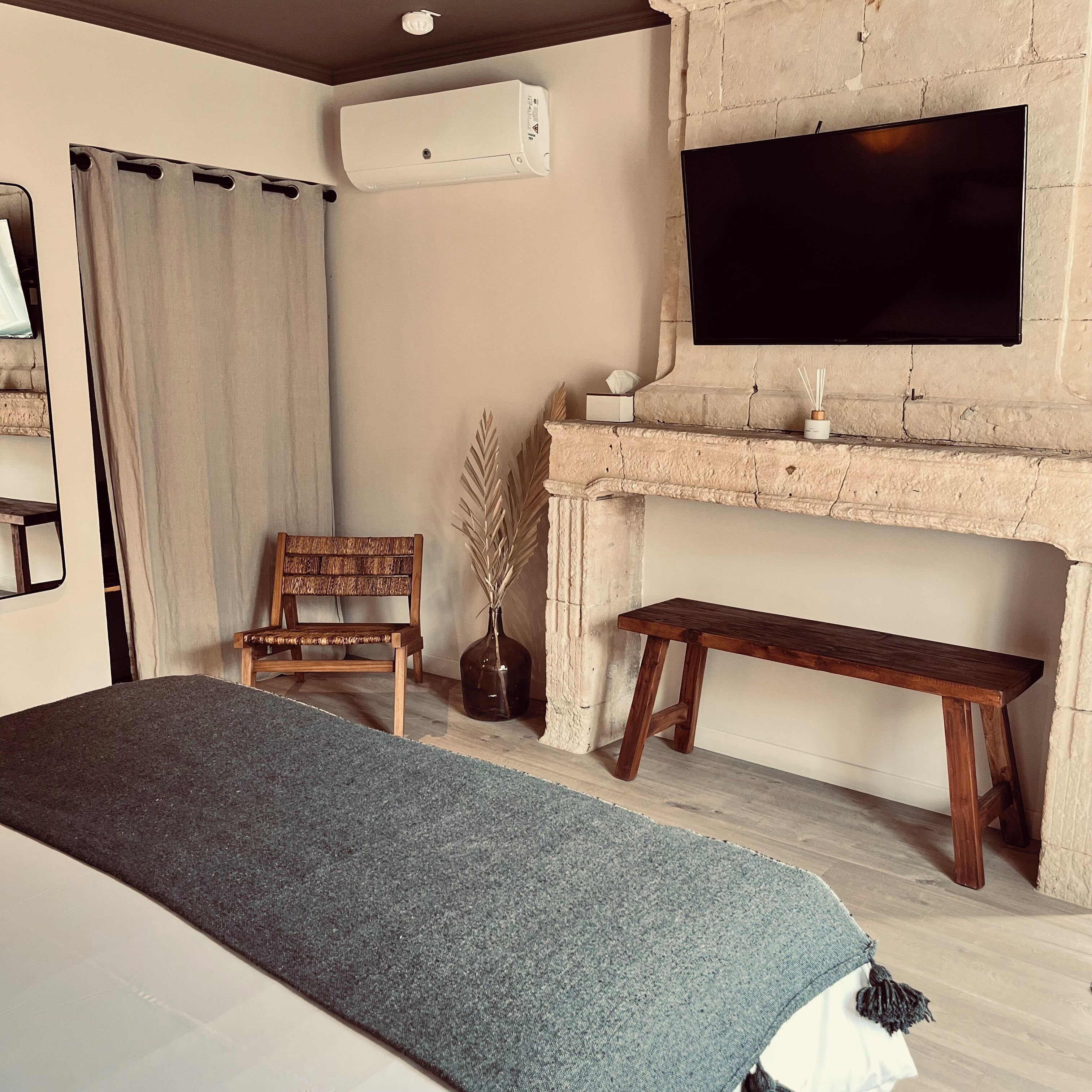 A modern living room with a large bed, a wooden chair, a bench, a fireplace, and a flat-screen TV mounted on the wall.