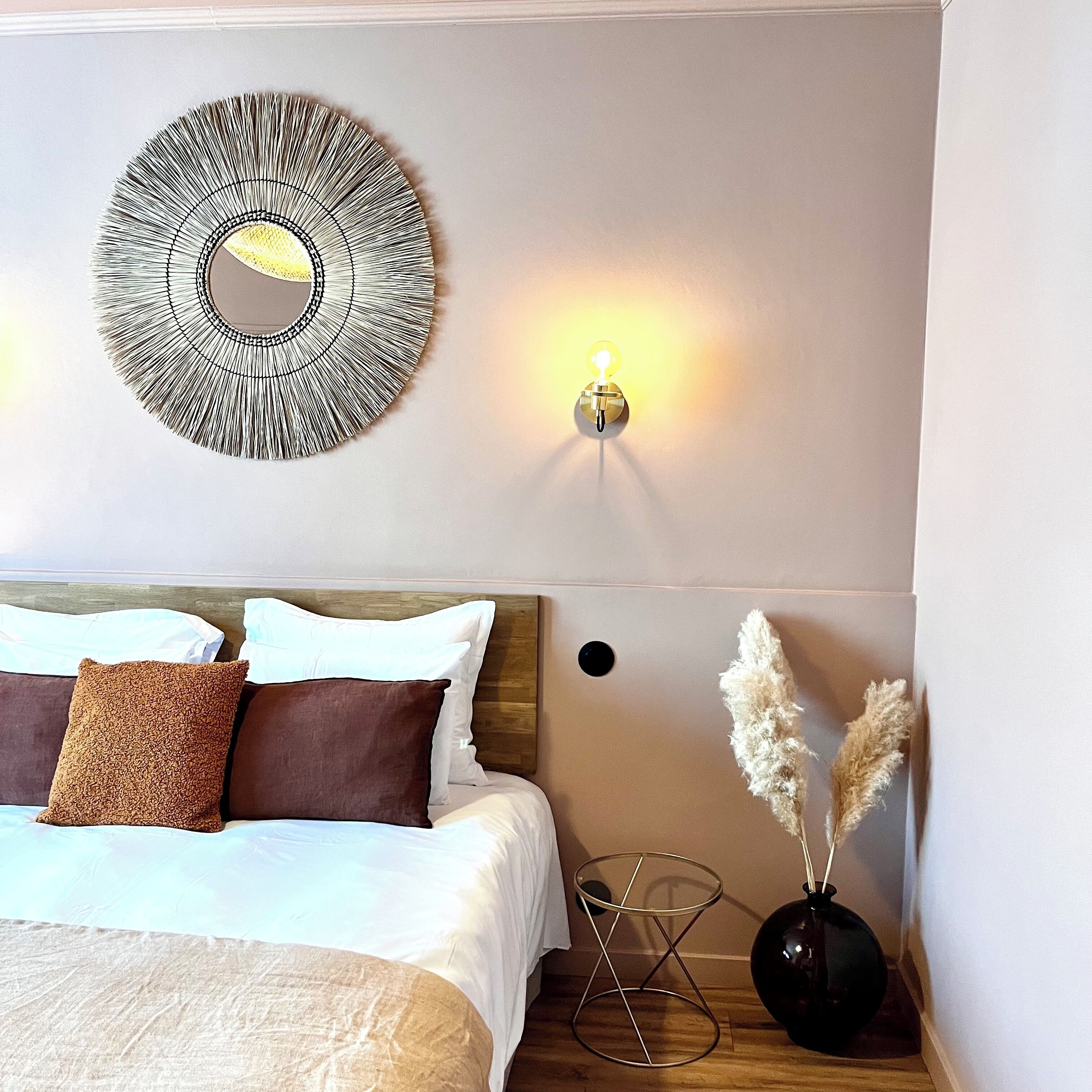 A neatly made bed with pillows, a wall-mounted mirror, a wall sconce, and a vase with pampas grass.