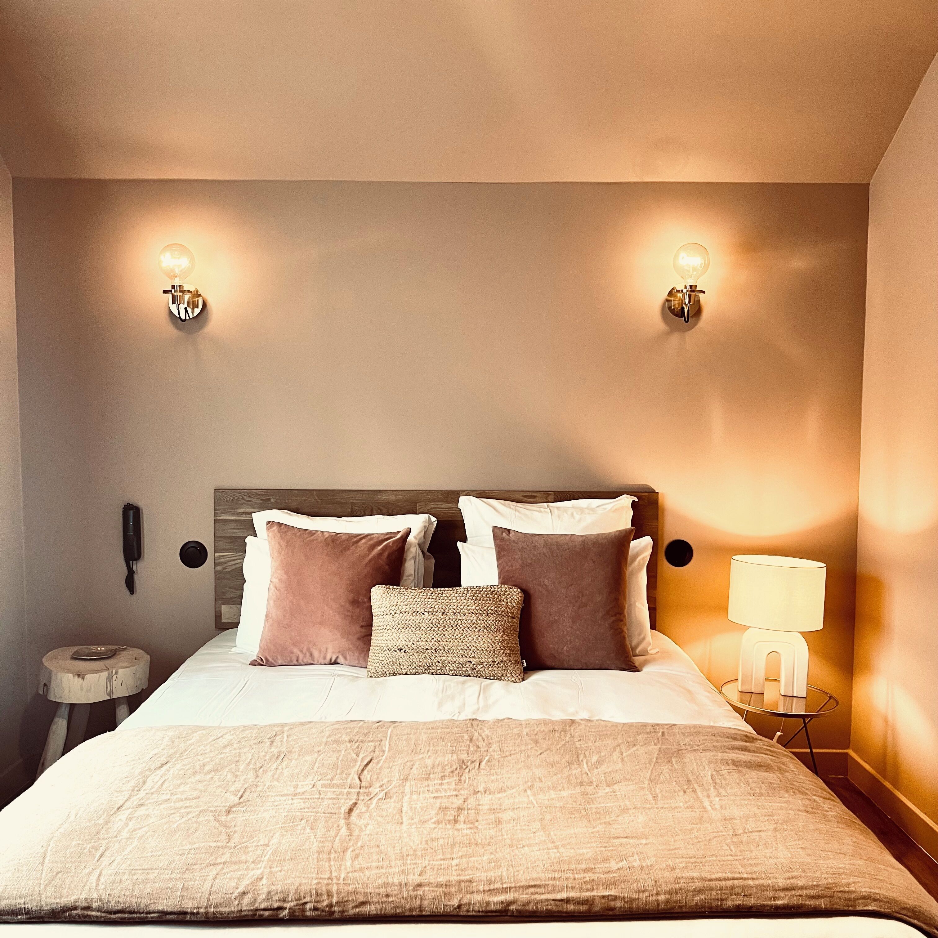 Una cama bien hecha con cabecera de madera, dos lámparas fijadas a la pared, una mesita de noche con lámpara y un teléfono.