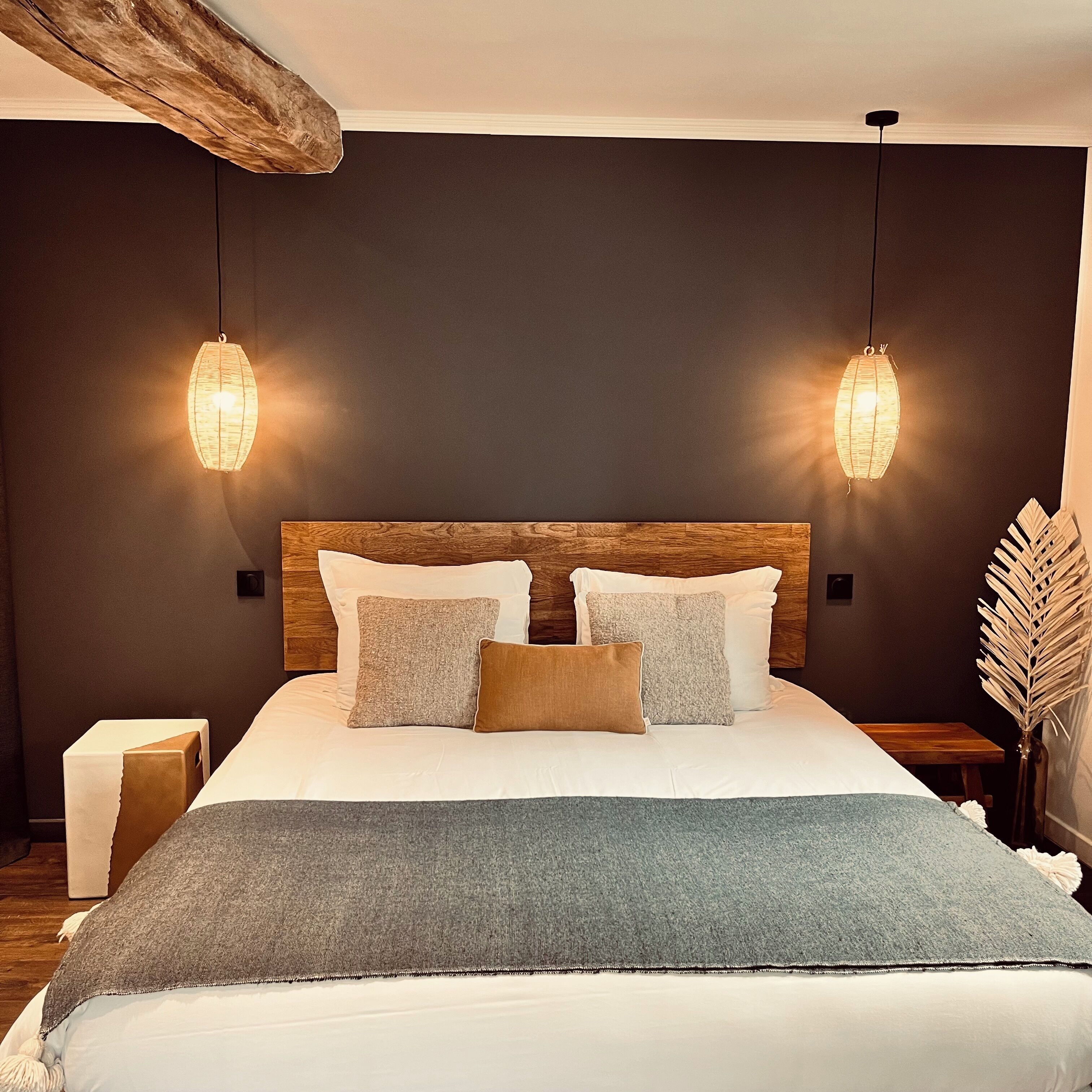 A bedroom with a wooden headboard, two bedside tables, a bed with white bedding and multiple pillows, and two hanging light fixtures.