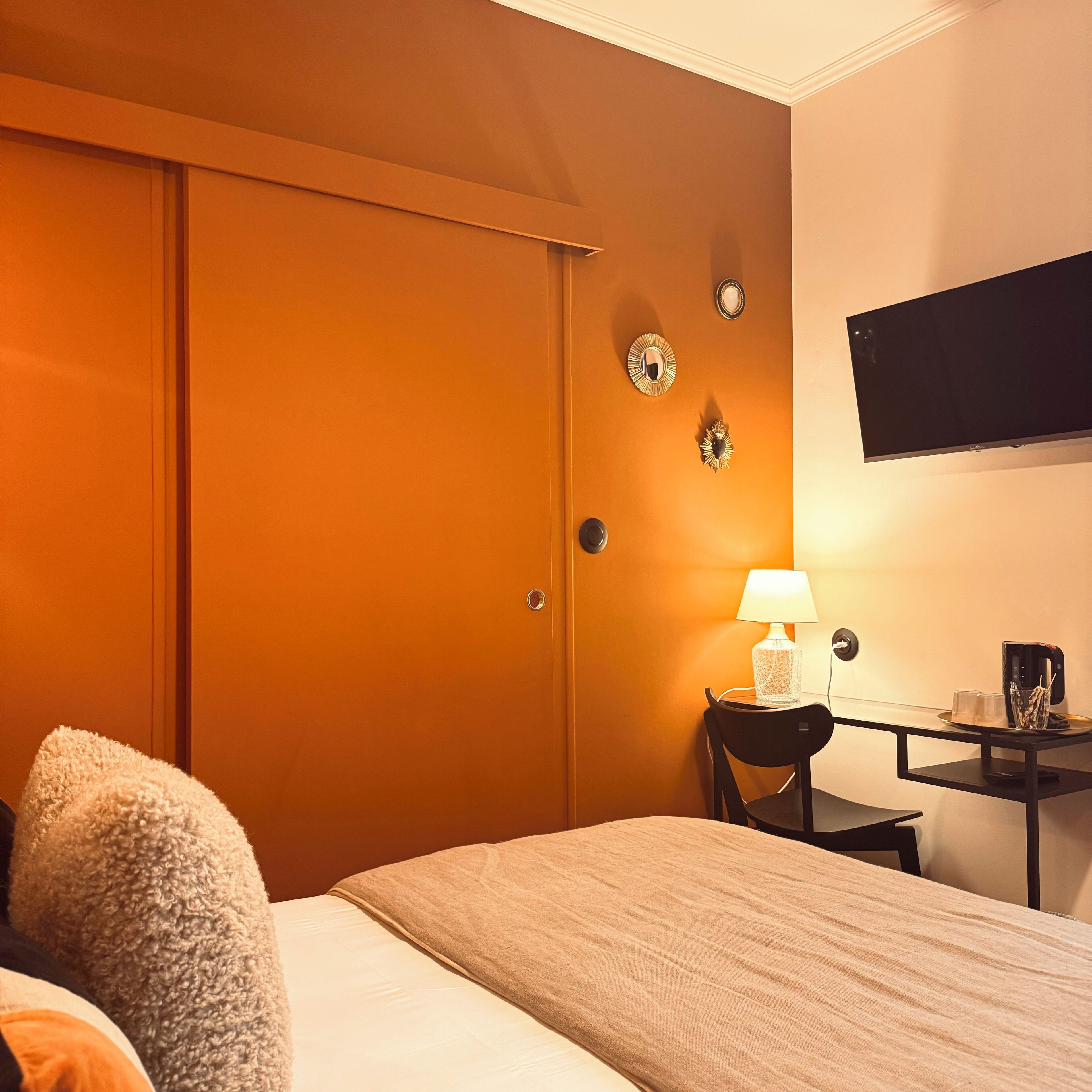 A hotel room with a bed, a desk with a lamp, a coffee maker, and a TV on the wall.