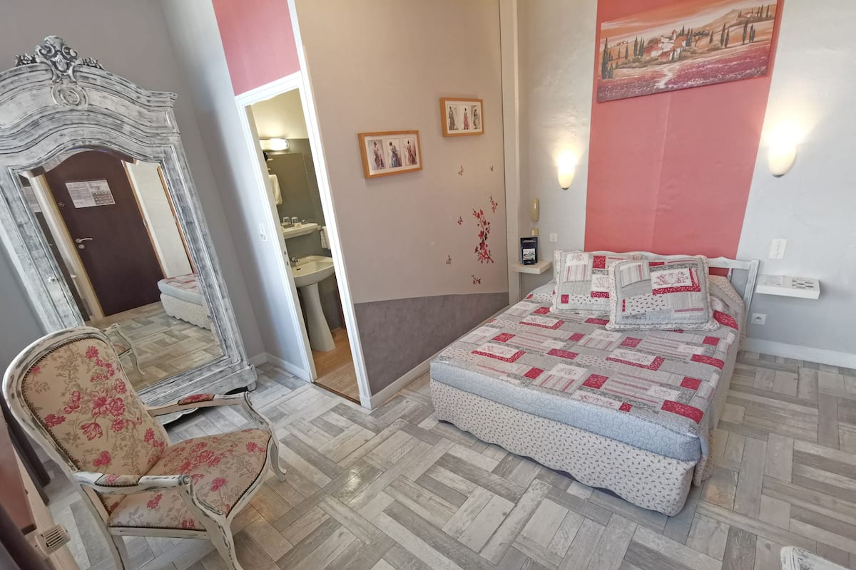 A bedroom with a bed, a chair, a mirror, and a bathroom visible through an open door.