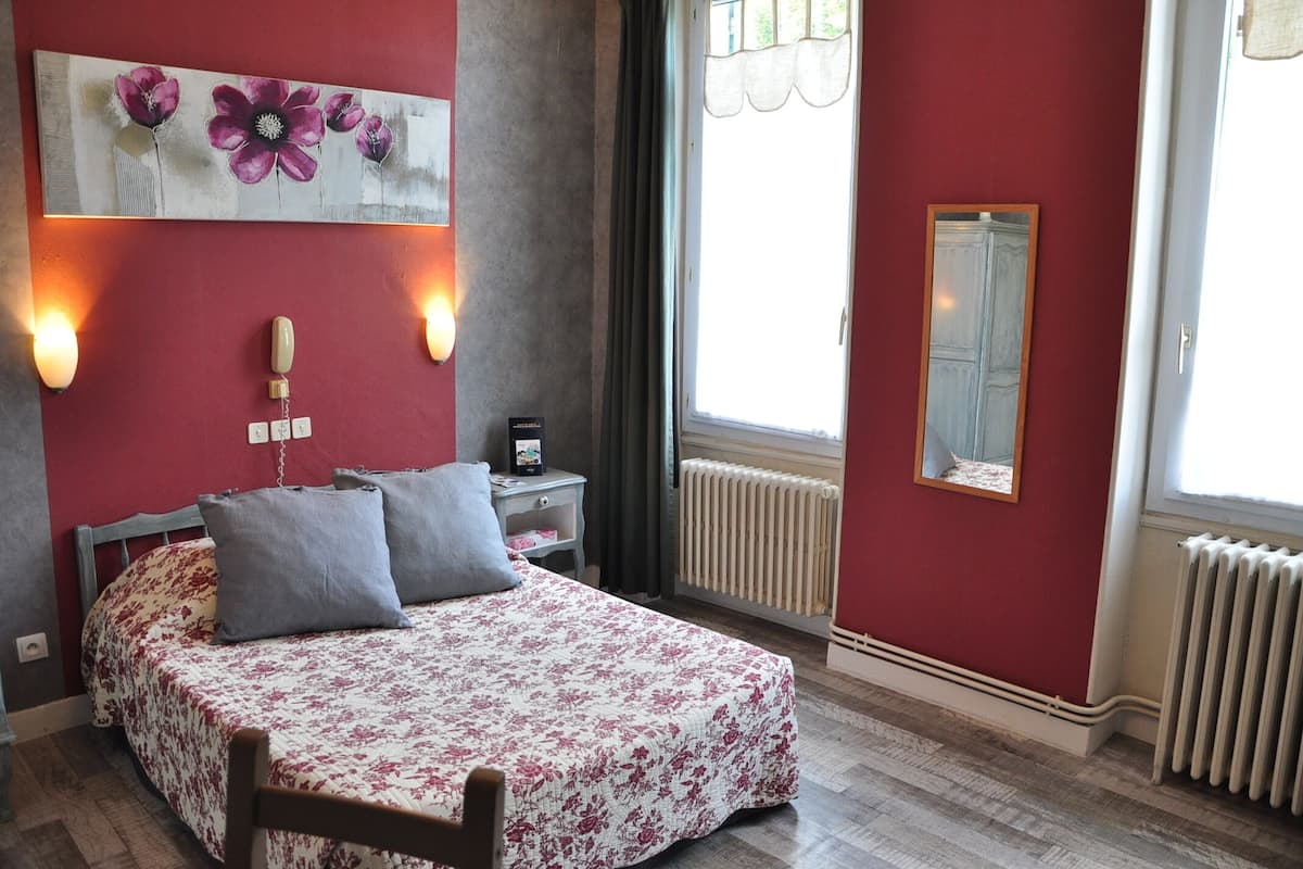 A bedroom with a bed, a nightstand, a wall-mounted light, a window with curtains, and a radiator.