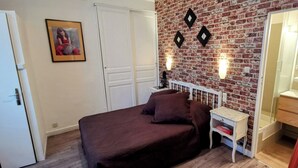 A bedroom with a brick wall, a bed with a brown bedspread, a nightstand, a mirror, and a striped chair. - Hôtel Select (Salon-de-Provence)