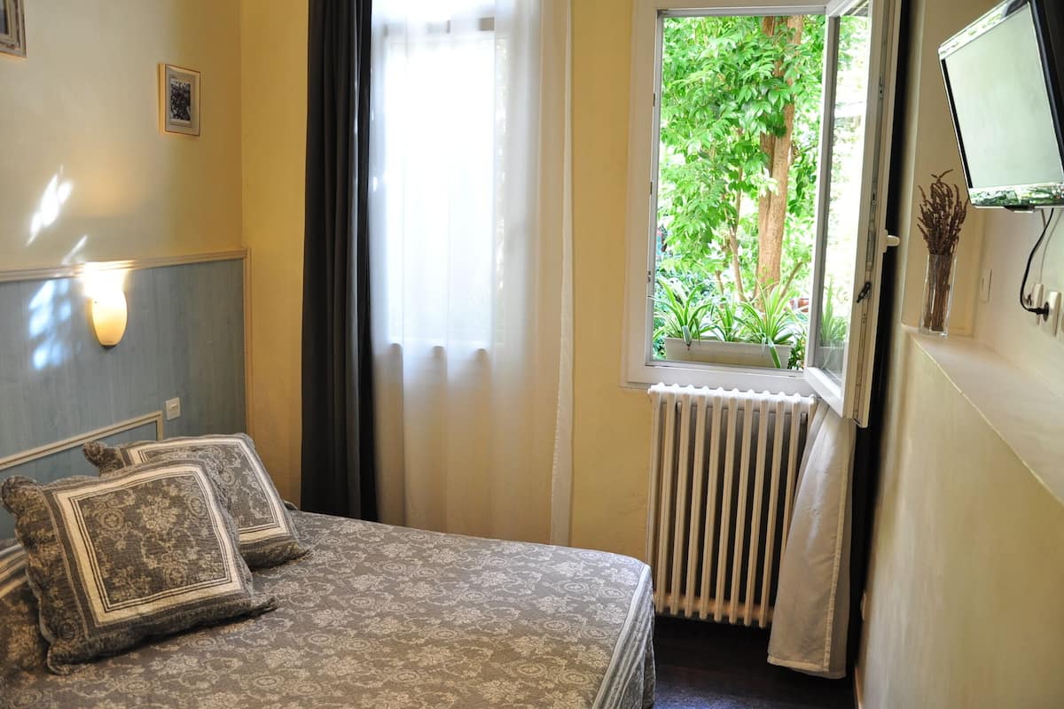 A bedroom with a bed, a window with a view of greenery, a radiator, and a wall-mounted TV.