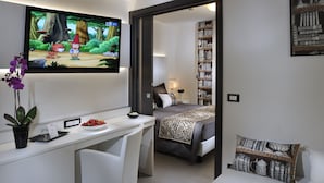 A modern hotel room with a desk, a bed with a zebra-patterned blanket, a bookshelf, and a TV displaying an animated show. - Hotel Rossini (Lignano Sabbiadoro)