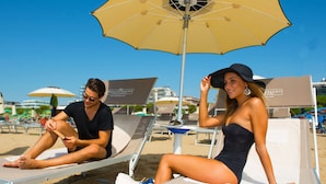 A beachside lounge area with sun loungers and umbrellas, a clear sky, and a sign indicating a hotel resort. - Hotel Rossini (Lignano Sabbiadoro)