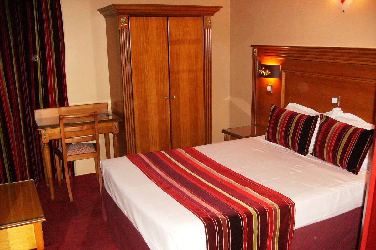A hotel room with a large bed, a wooden desk, a wardrobe, and a nightstand with a lamp.