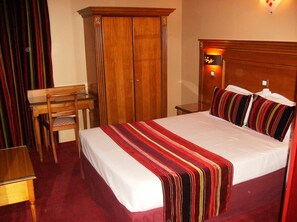 A hotel room with a large bed, a wooden desk, a wardrobe, and a nightstand with a lamp. - Hôtel du Val de Seine (Mantes-la-Jolie)