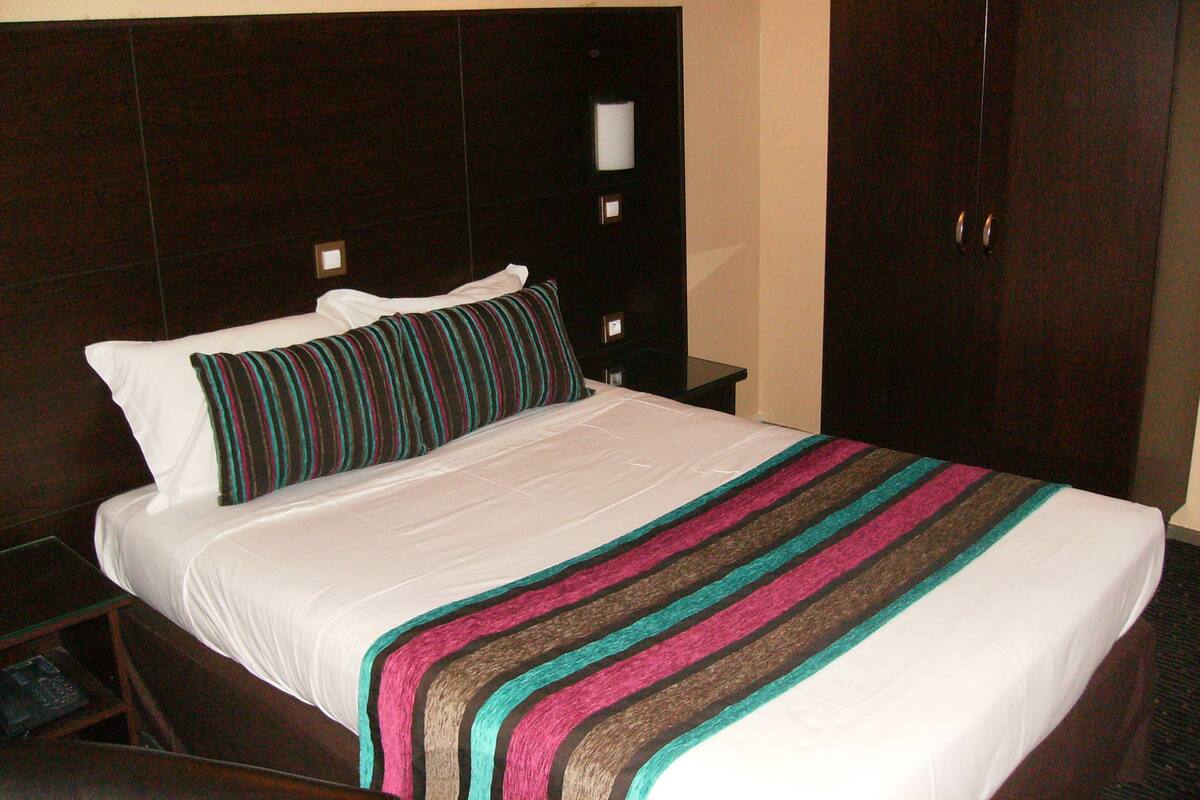A hotel room with a large bed, a dark wooden headboard, a nightstand with a lamp, and a wardrobe.