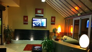 A modern lobby with a reception desk, a flat-screen TV mounted on the wall, a water feature, and several potted plants. - Hôtel du Val de Seine (Mantes-la-Jolie)