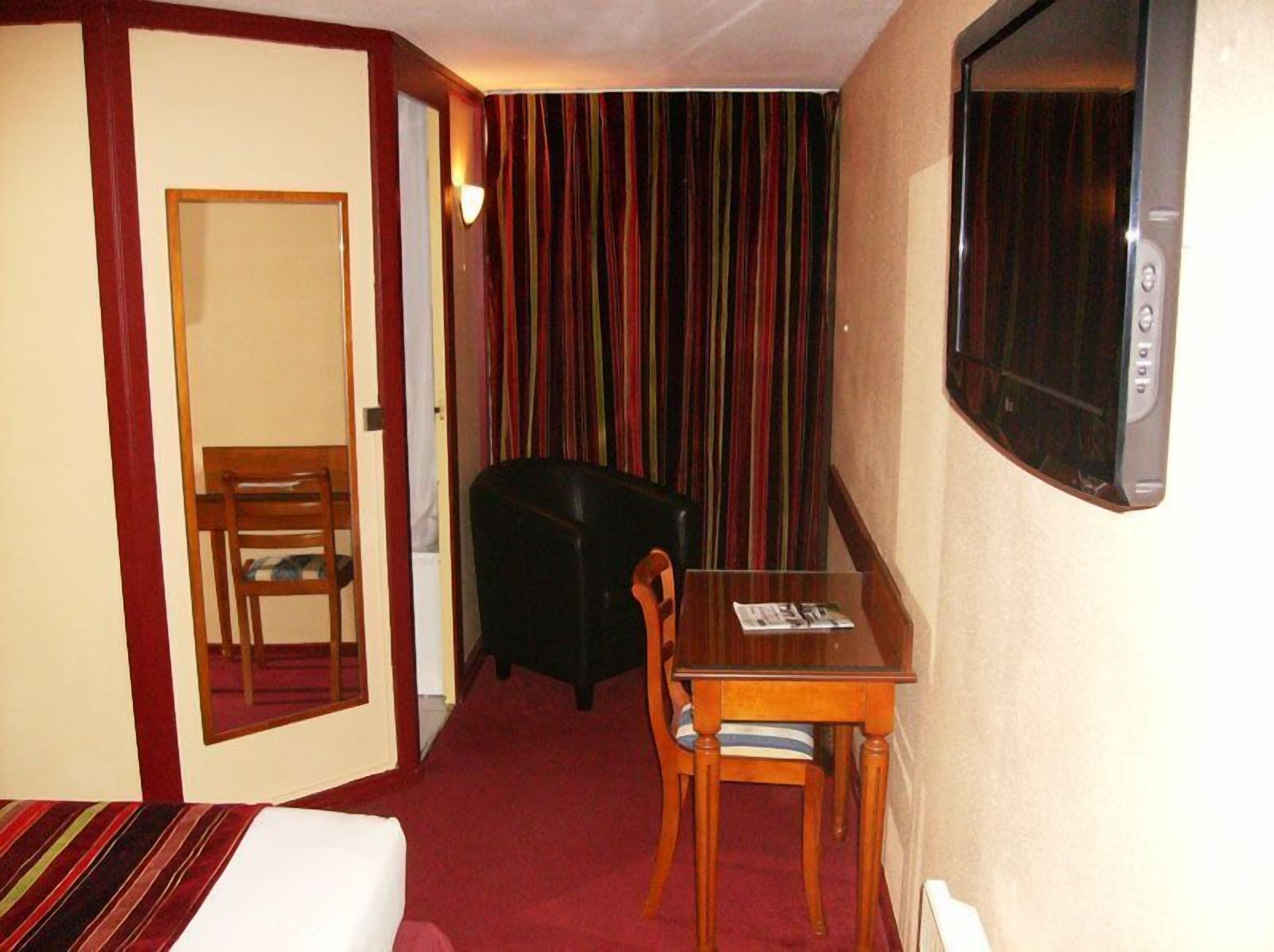 Una habitación de hotel con una cama, una mesa de madera con dos sillas, un sillón negro, una cortina a rayas y un televisor fijado a la pared.