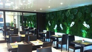 A restaurant with tables and chairs, a green wall with plants, and a ceiling with lights. - Hôtel du Val de Seine (Mantes-la-Jolie)