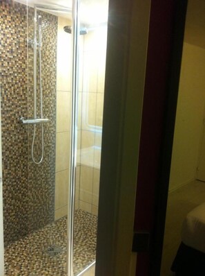 A shower area with a tiled wall and floor, a glass door, and a visible showerhead. - Hôtel du Val de Seine (Mantes-la-Jolie)