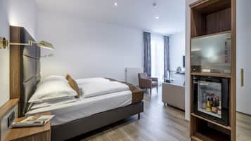 A modern hotel room with a large bed, a wooden side table, a built-in minibar, and a seating area with a chair and a small table.