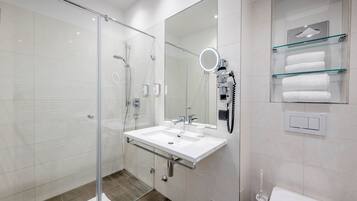 A modern bathroom with a glass shower enclosure, a white sink with a mirror above it, and a glass shelf with towels.