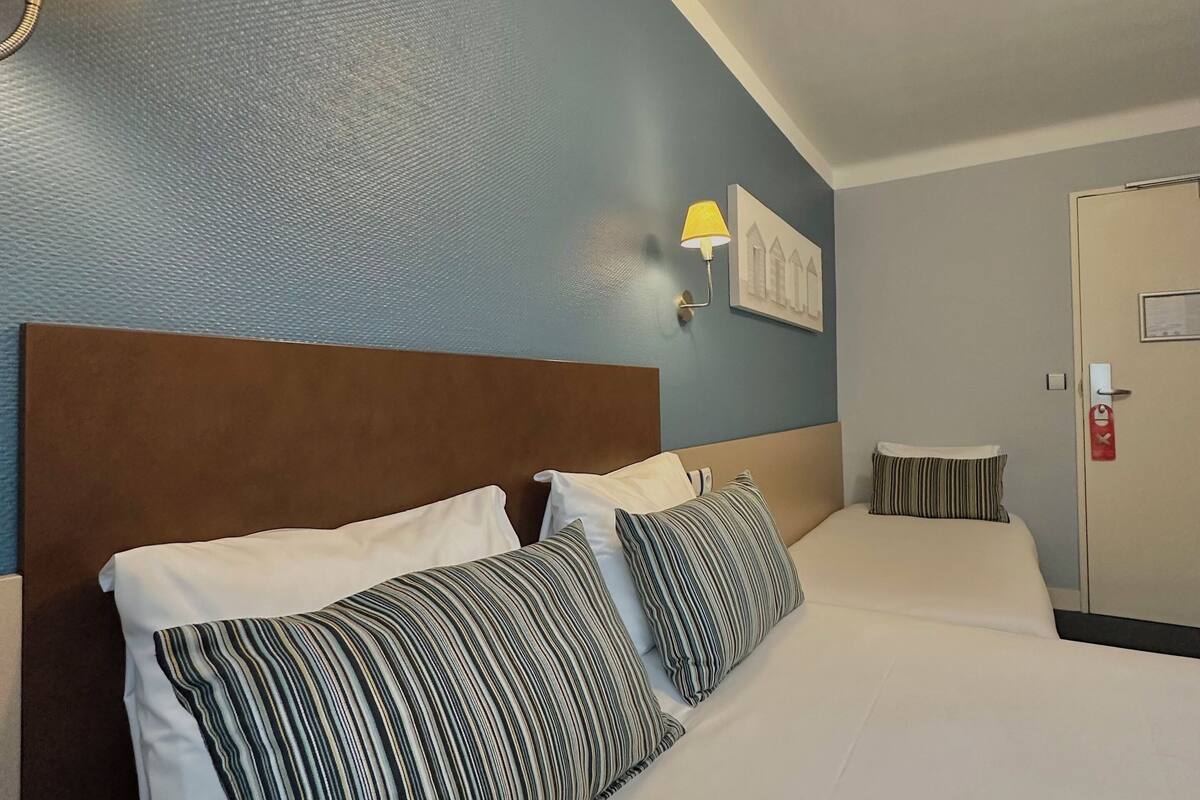 Une chambre d’hôtel avec un grand lit, une tête de lit, une lampe fixée au mur et une porte équipée d’un détecteur d’incendie.