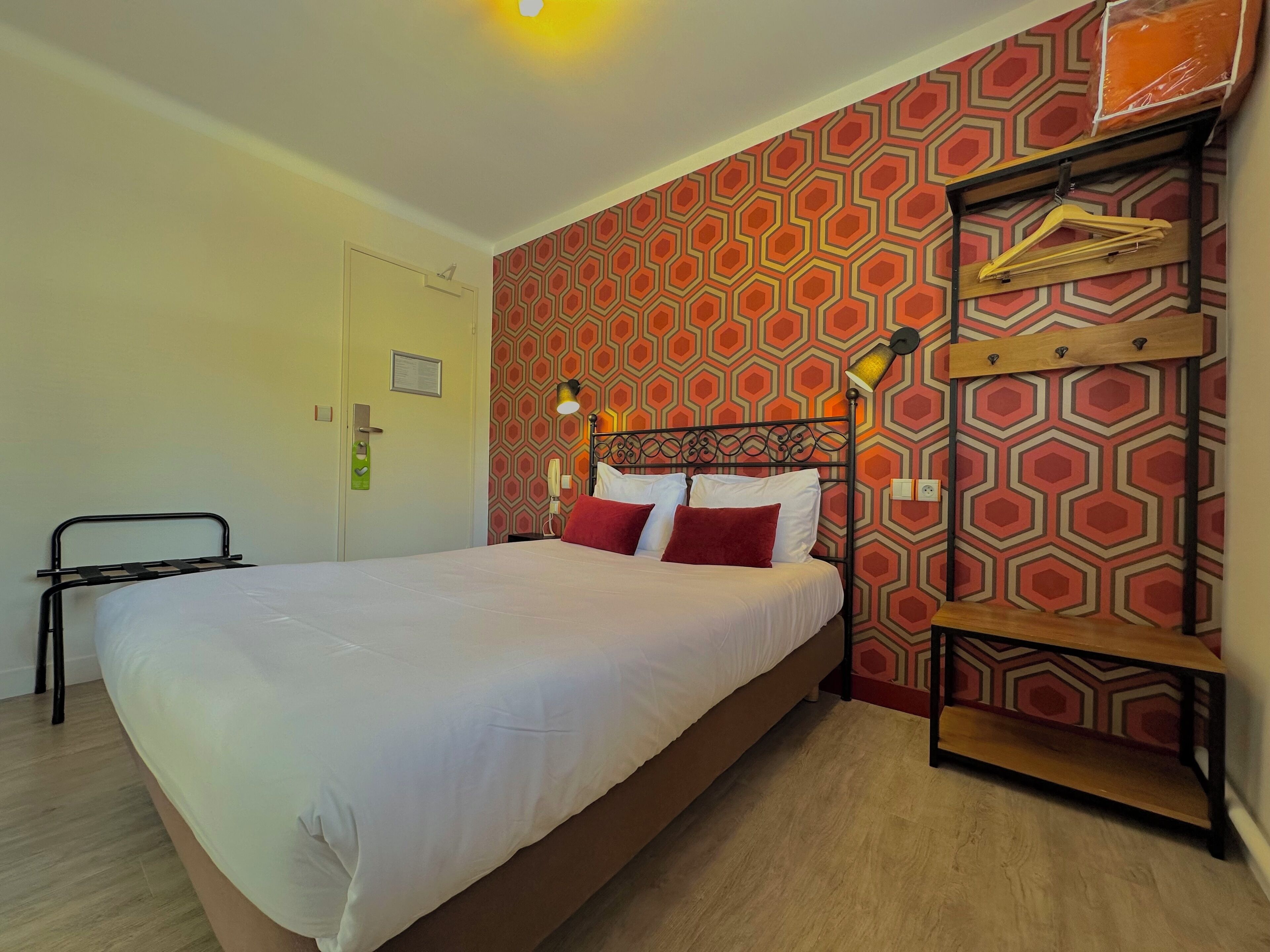 A hotel room with a large bed, a bedside table with a lamp, a wardrobe with hanging clothes, and a door with a handle.