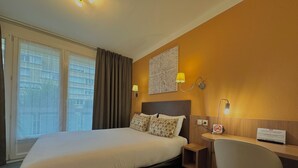 A hotel room with a large bed, a desk with a chair, a lamp, and a view of the city through the window. - Hôtel de Caen (Caen)
