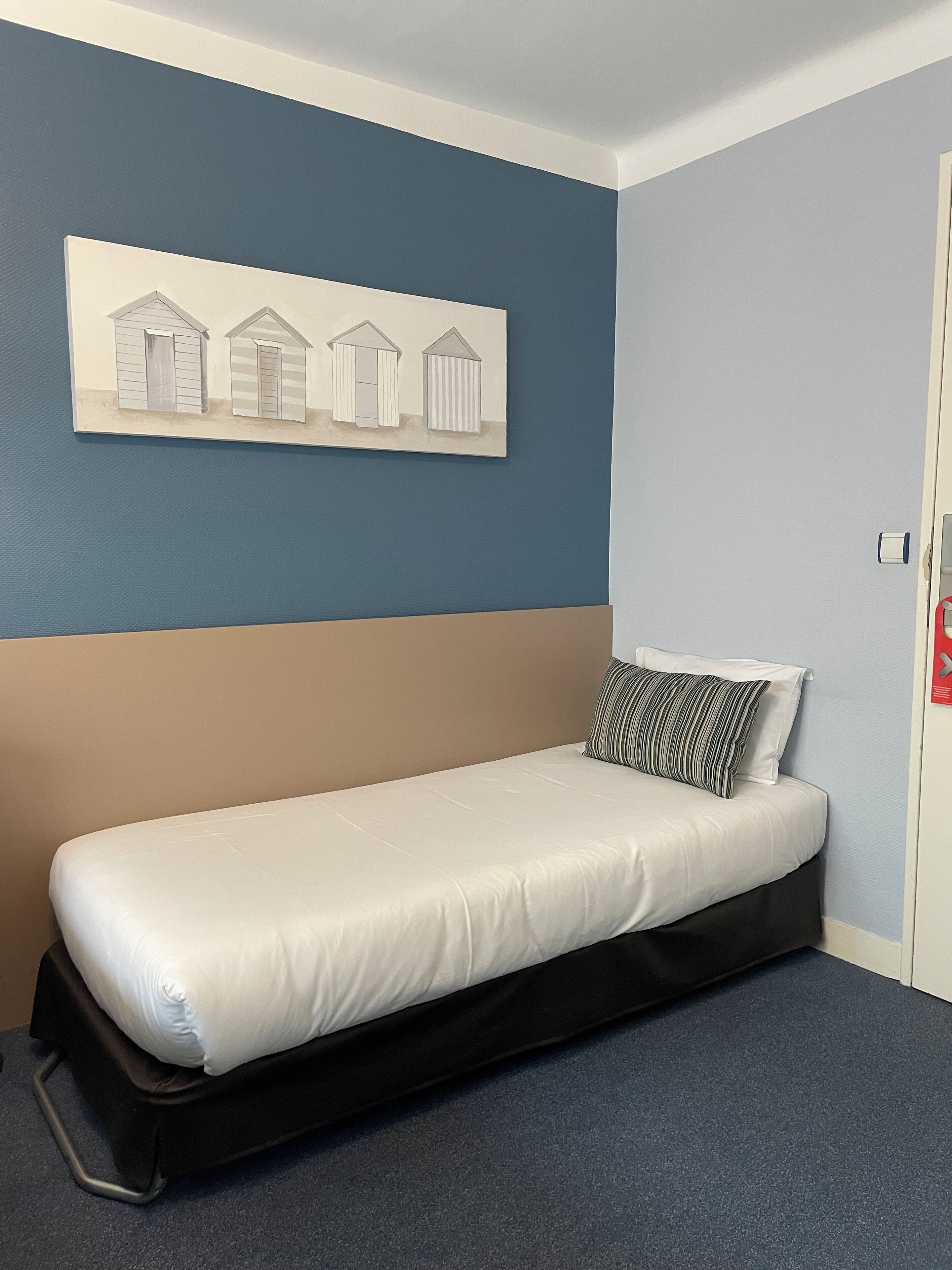 Un lit simple avec un matelas blanc et un cadre noir, un oreiller à rayures et un tableau accroché au mur représentant trois cabanes de plage.