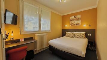 A hotel room with a bed, a desk with a chair, a television, a radiator, and a window with blinds.