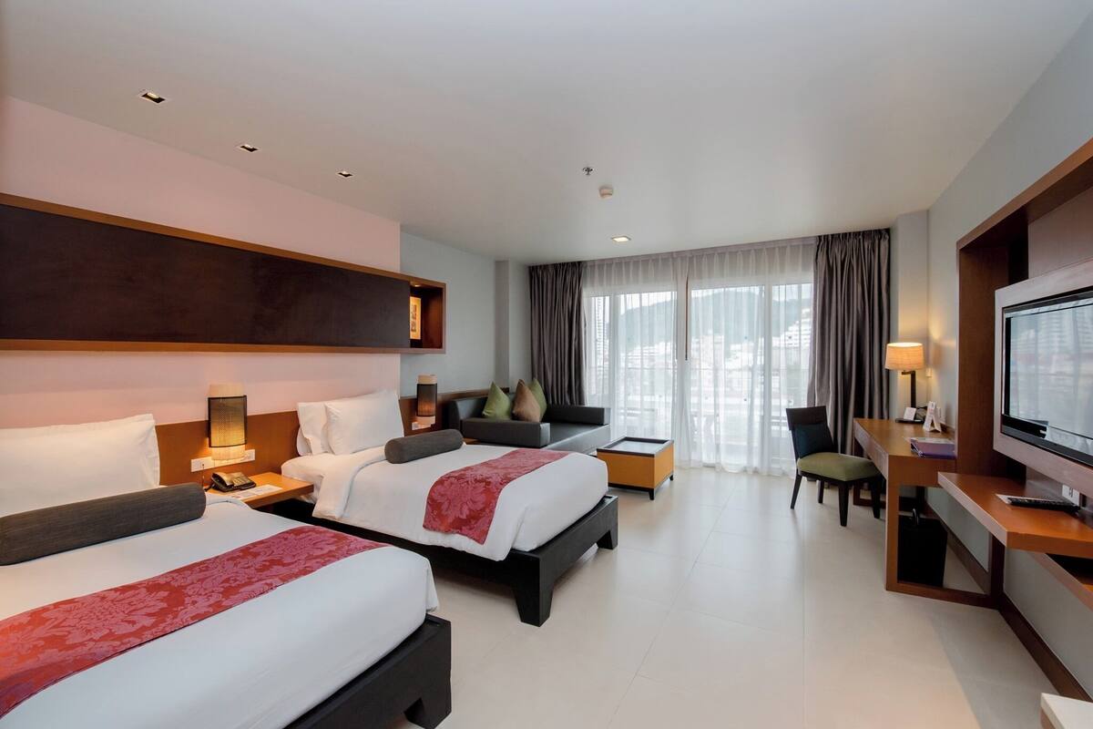A hotel room with two beds, a desk, a TV, and a large window with curtains.