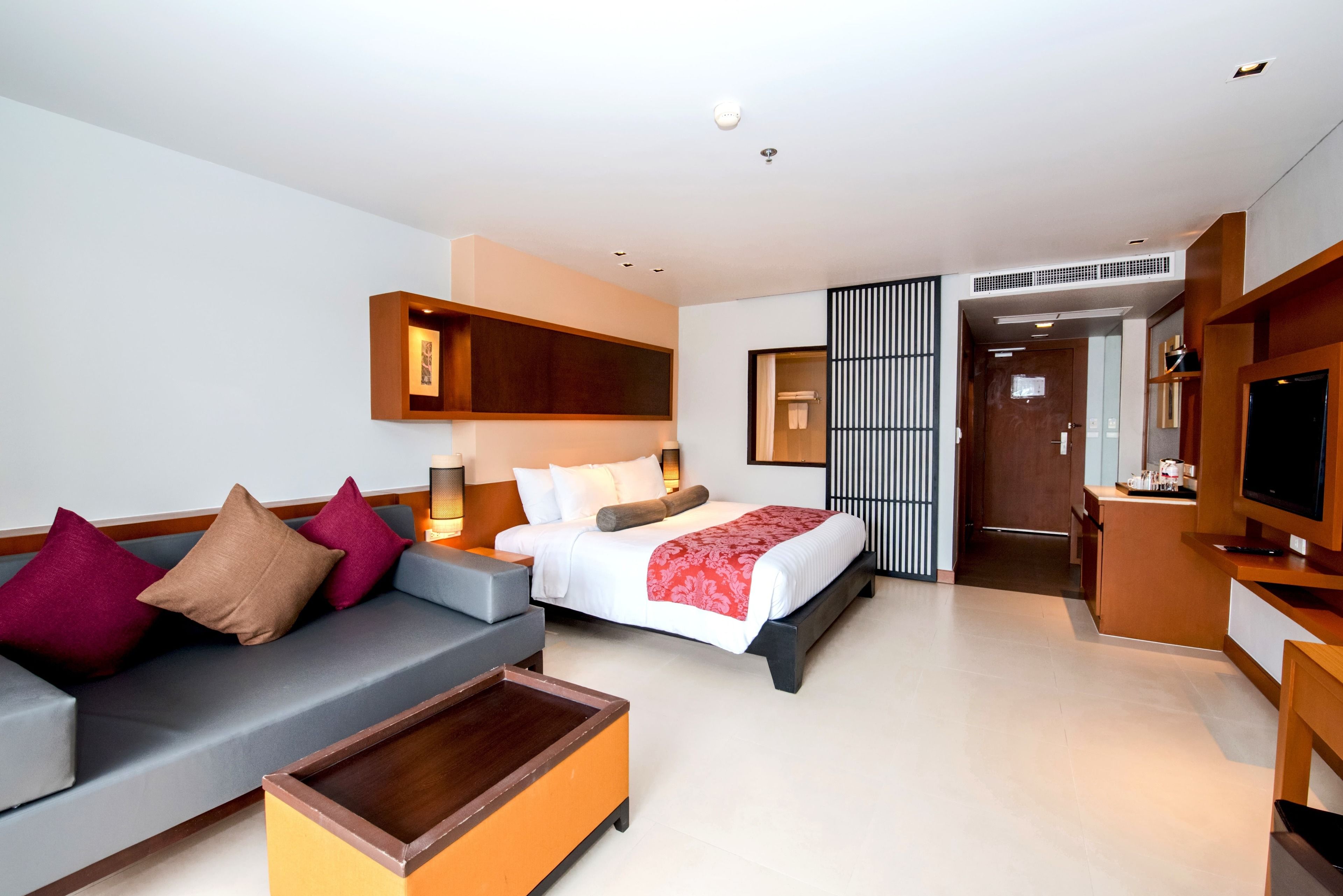 A modern hotel room with a sofa, a bed with a red blanket, a TV, and a small table.