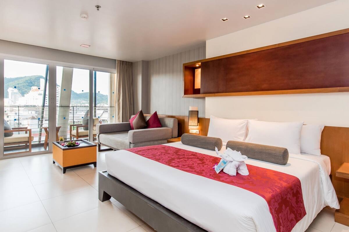 A modern hotel room with a large bed, a sofa, a small table, and a view of the city.