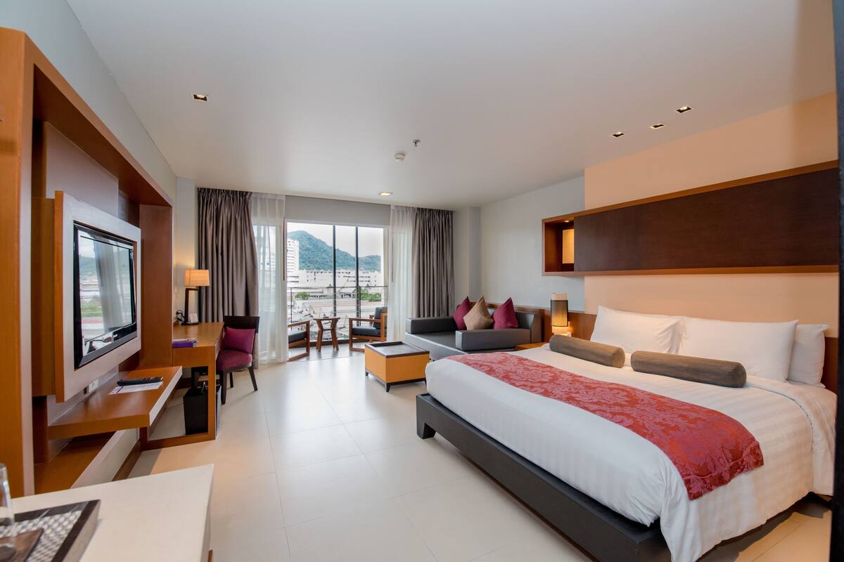 A modern hotel room with a large bed, a TV, a desk, and a seating area with a view of the city.