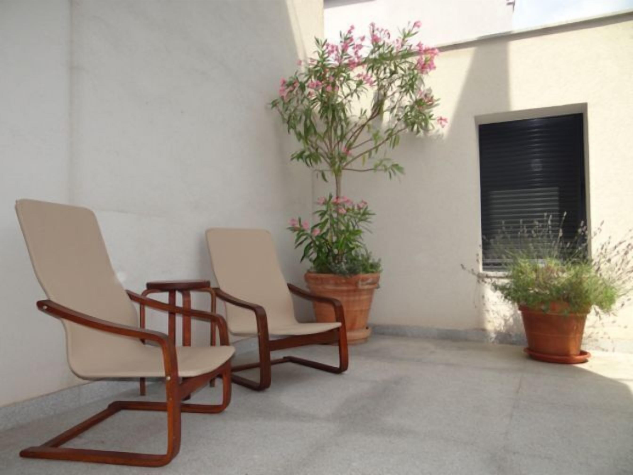 Two wooden chairs with beige cushions, a potted plant with pink flowers, and a window with a closed shutter.