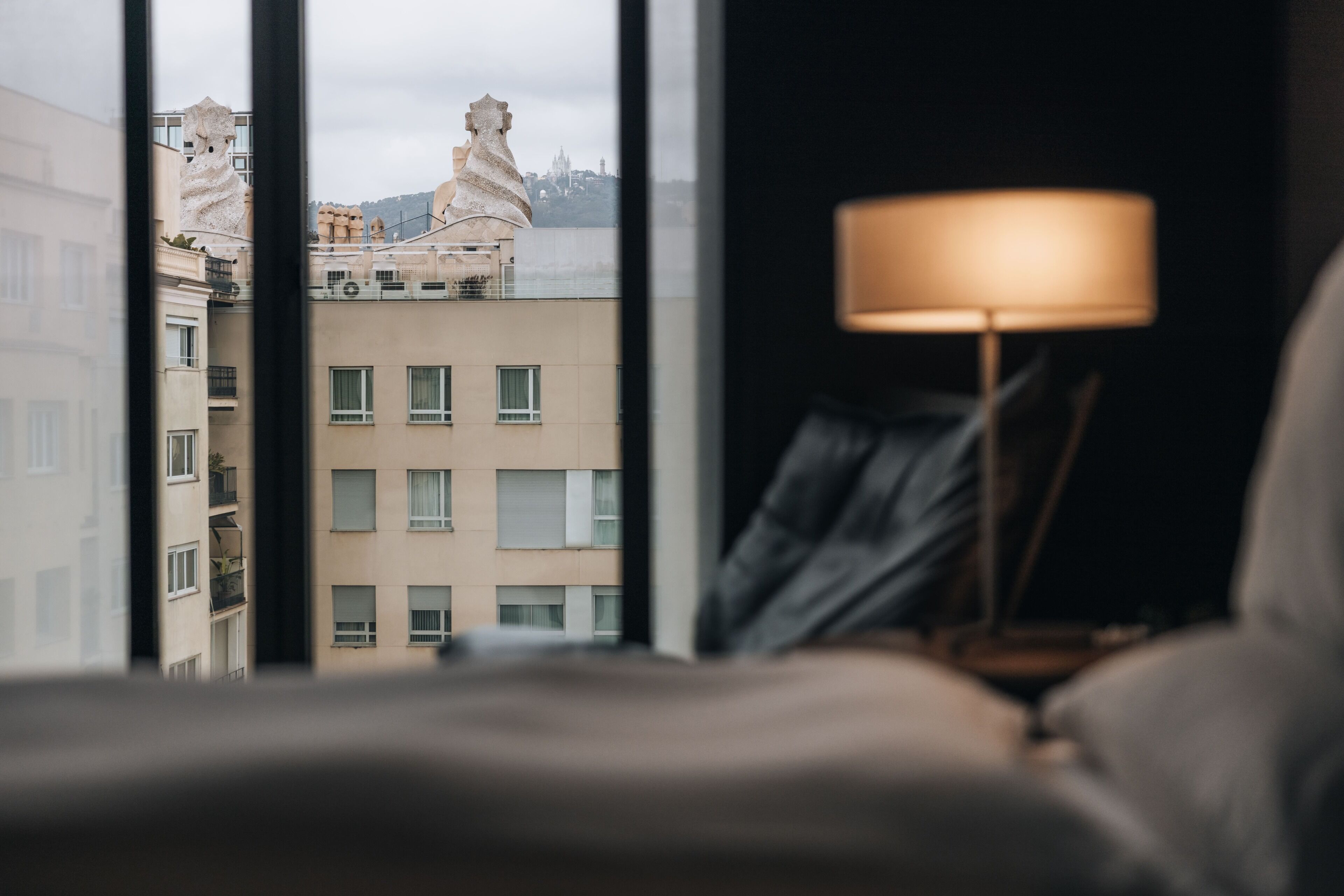 Una habitación con vistas a edificios a través de una ventana, una lámpara sobre una mesita de noche y una cama.